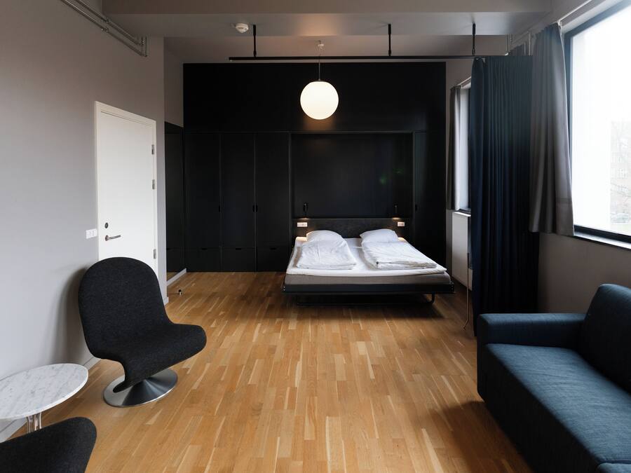 A modern hotel room with a wooden floor, a bed with white linens, a dark blue sofa, a round black chair, and a small white table.