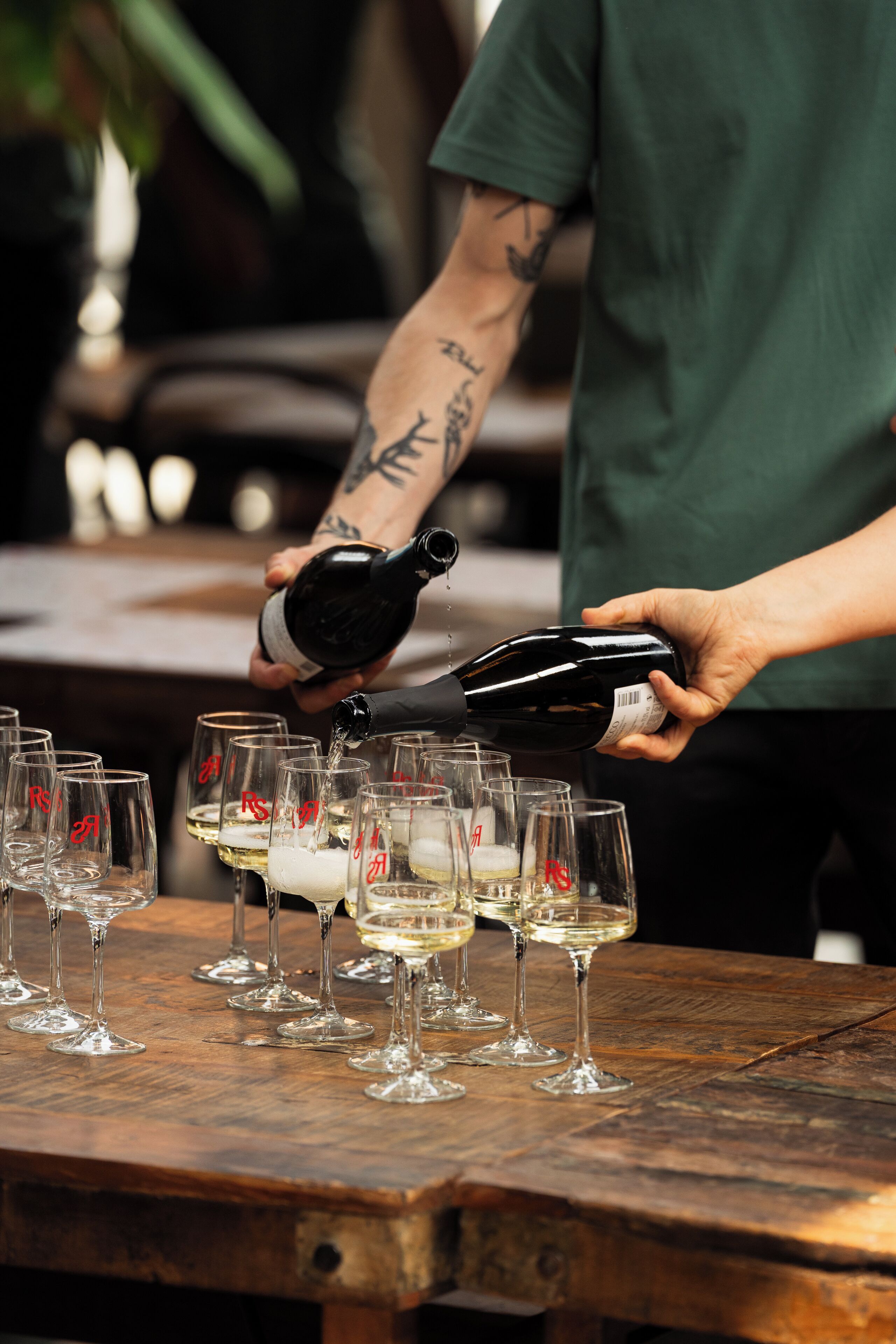 A person pouring a liquid from a bottle into several glasses on a wooden table.