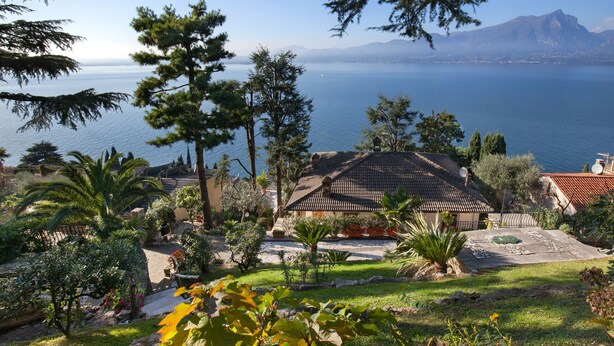A house with a tiled roof, surrounded by lush greenery and a view of a large body of water and mountains in the distance. - Villa Vista Lago With Pool (Torri del Benaco)