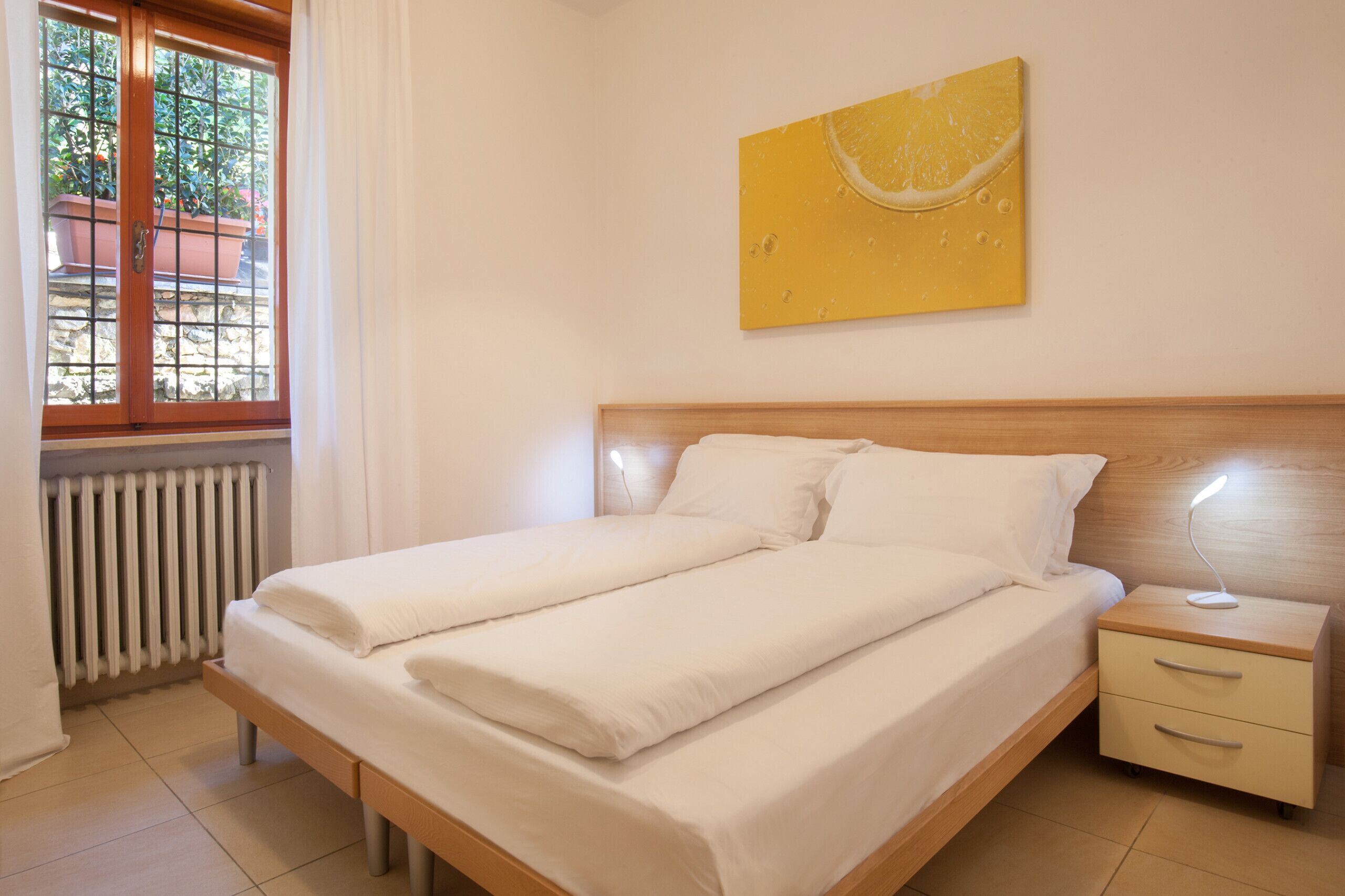 A double bed with white linens, a wooden headboard, a bedside table with a lamp, and a window with curtains.