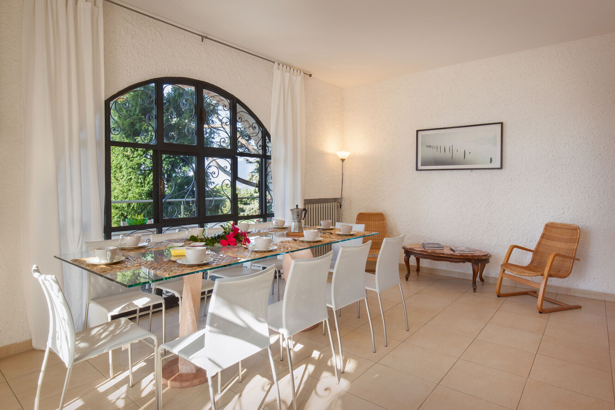 A dining area with a glass table, white chairs, a wooden side table, and a large window with curtains.