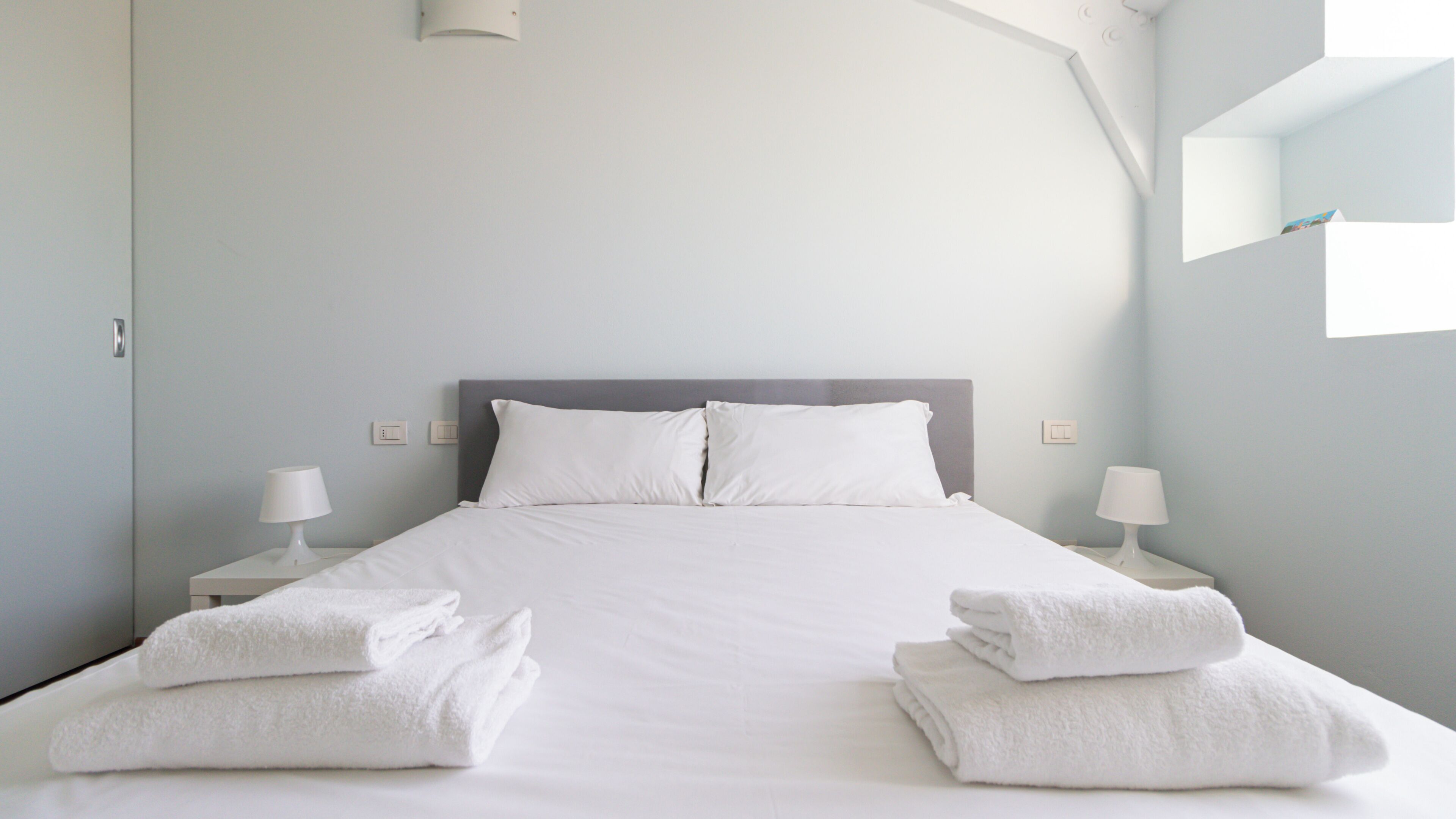 Una cama bien tendida con sábanas y almohadas blancas, dos lámparas de noche y una pila de toallas dobladas a cada lado.