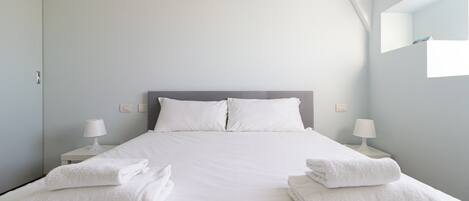 Una cama bien tendida con sábanas y almohadas blancas, dos lámparas de noche y una pila de toallas dobladas a cada lado.
