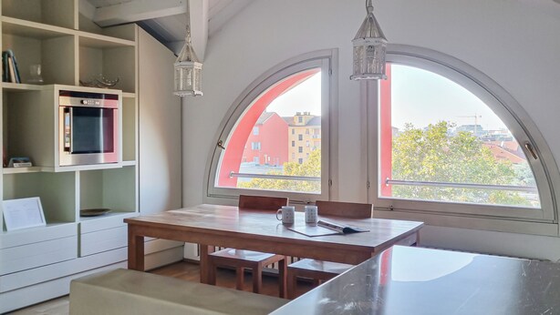 Cocina moderna con comedor, ventanas arqueadas amplias y vista a edificios y árboles.