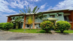 Un edificio de varios pisos con techo verde, rodeado de jardines cuidados y una palmera.