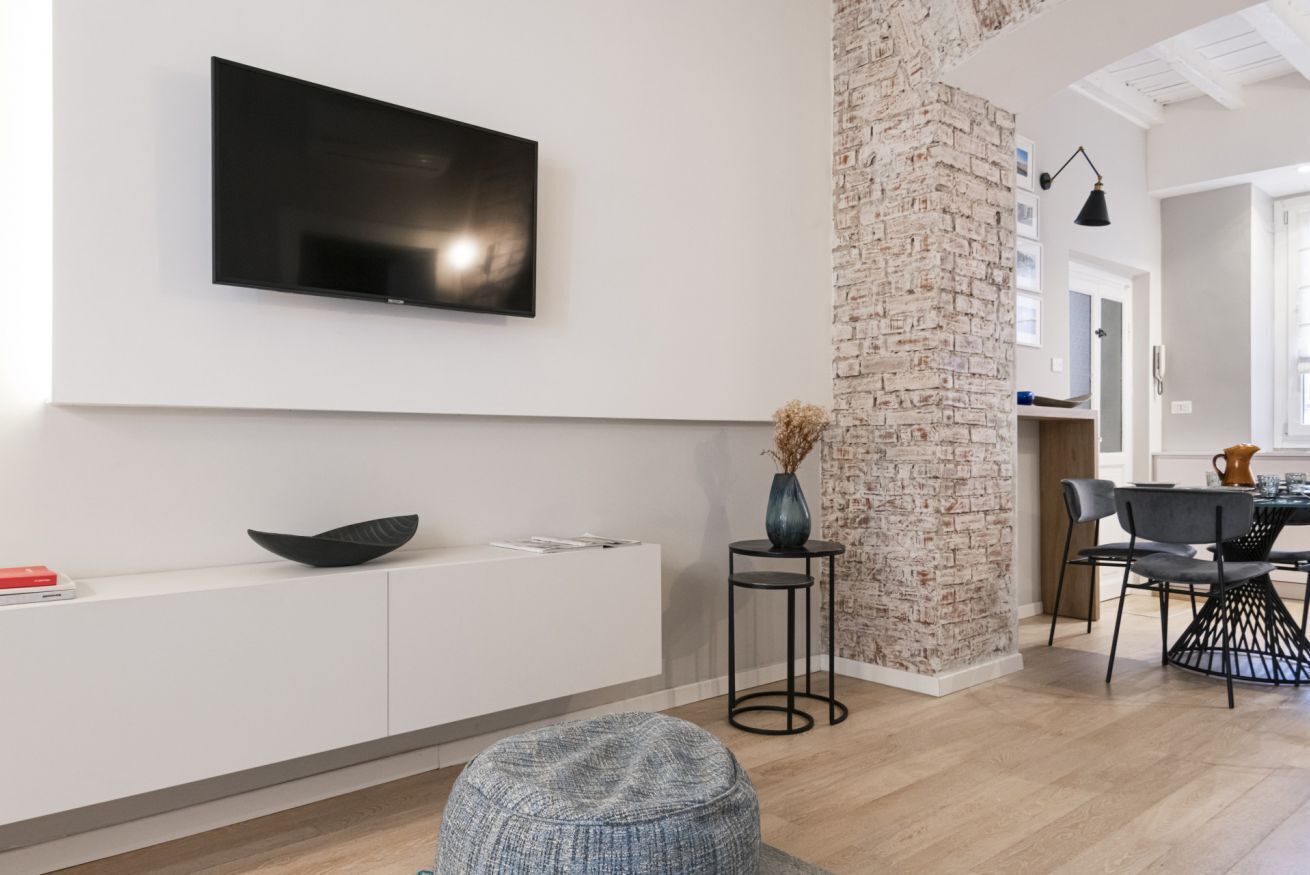 Ein modernes Wohnzimmer mit einem flachbildfernseher an der Wand, einem weißen Konsolentisch mit einer schwarzen Schüssel, einem grauen Pouf und einer Ziegelwand als Blickfang.