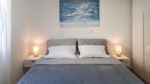 Un lit bien fait, recouvert d’une courtepointe grise matelassée, avec deux oreillers et deux lampes de chevet posées sur des tables de chevet assorties. Un tableau est accroché au-dessus du lit.