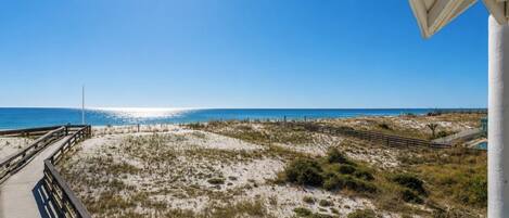 Vista de la playa con un muelle de madera que lleva al agua, dunas de arena y un cielo azul despejado.