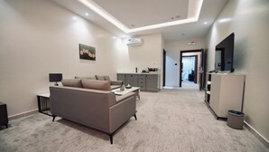 A modern living room with a sofa, a coffee table, and a TV. There is a kitchen area with cabinets and a microwave. The room has a carpeted floor and recessed ceiling lighting. - qimampark albaha7 (Al Baha)