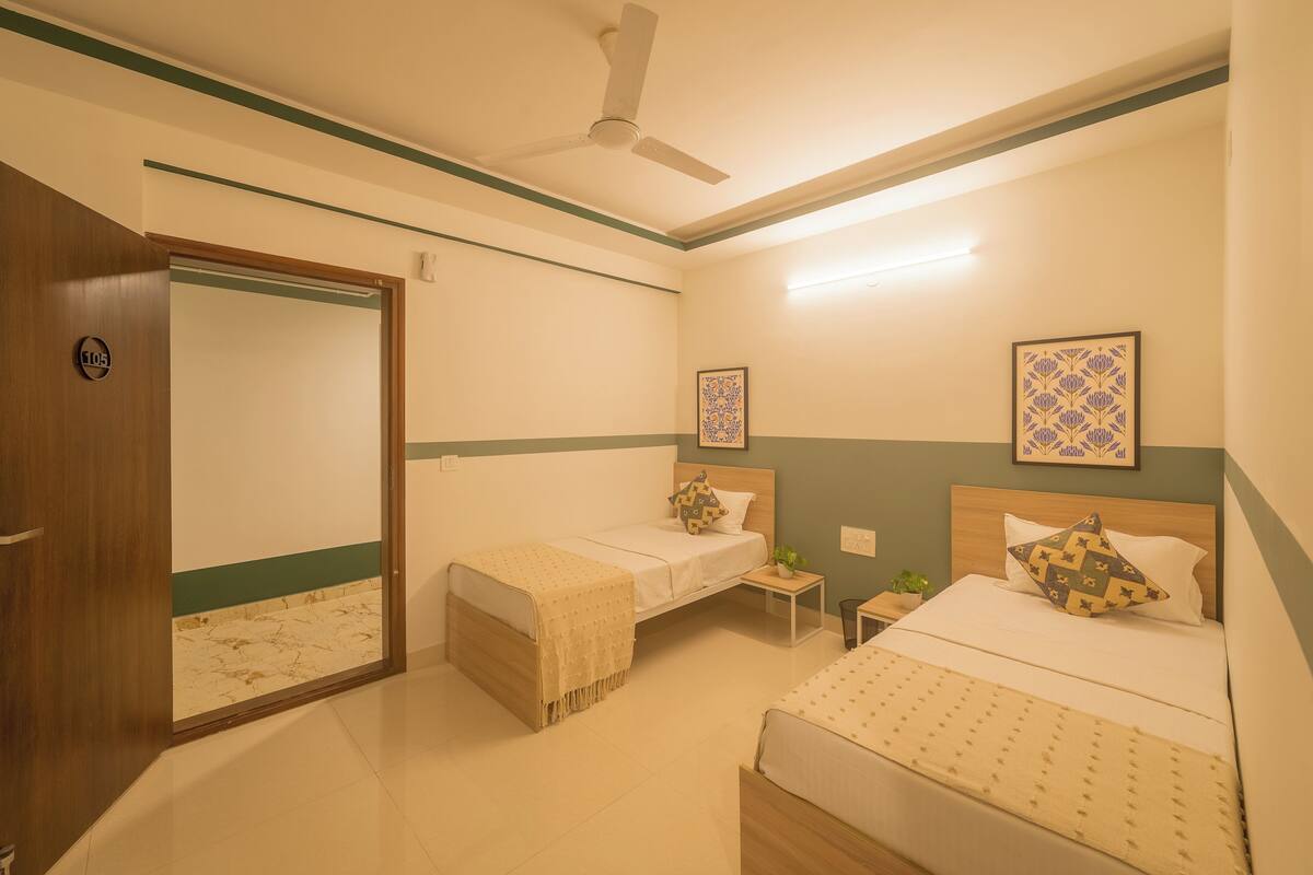 A hotel room with two beds, a ceiling fan, a nightstand with a lamp, and a mirror.
