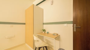 Una habitación pequeña y limpia con una puerta de madera, una silla blanca, un escritorio pequeño con una planta y un libro, y un cubo de basura.