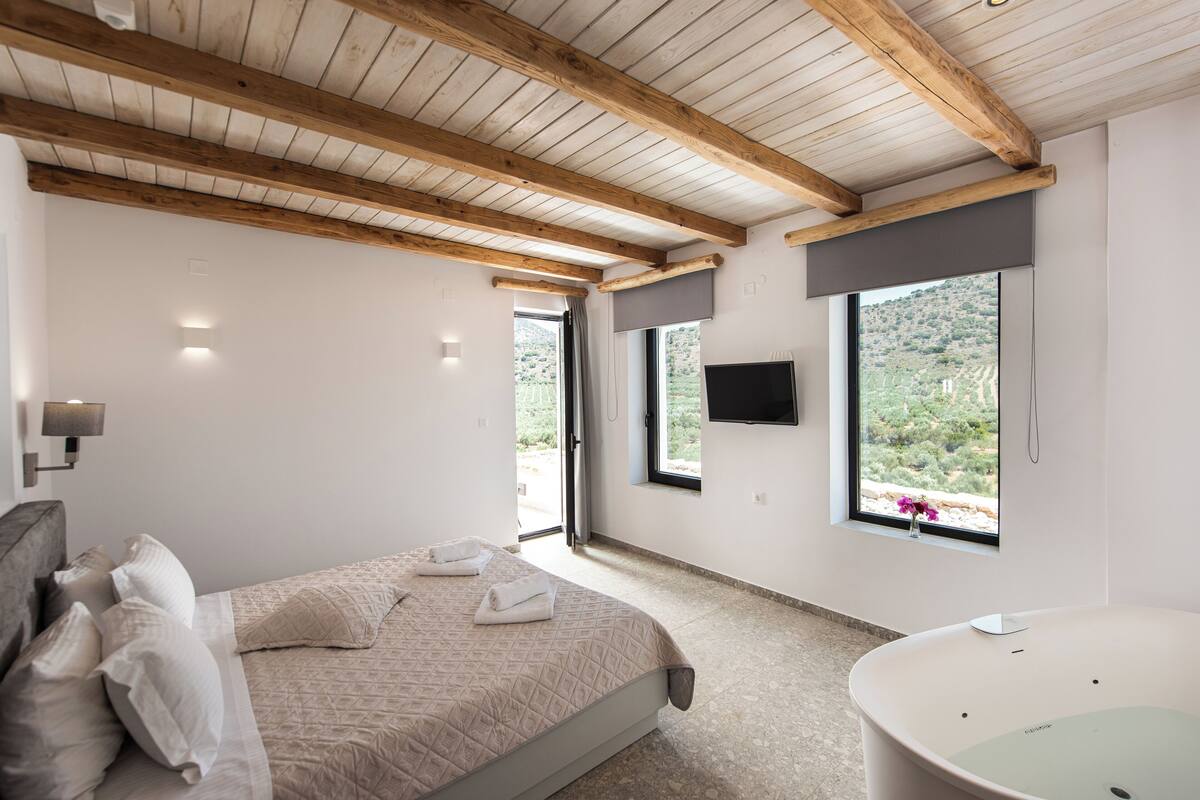 Een slaapkamer met een bed, een badkuip en twee ramen met uitzicht op een heuvelachtig landschap.