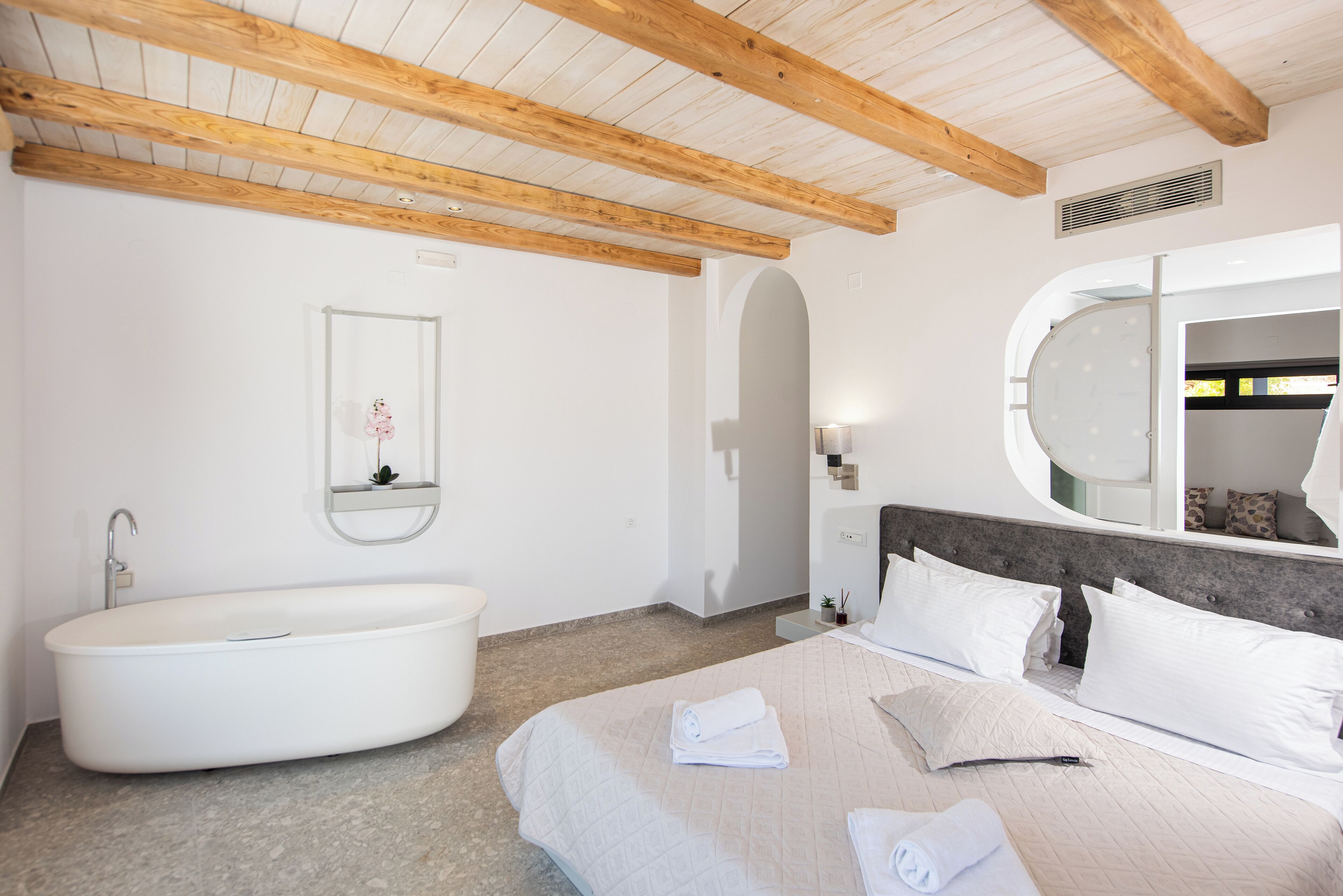 A bedroom with a large bed, a built-in bathtub, a round mirror, and a wooden ceiling.