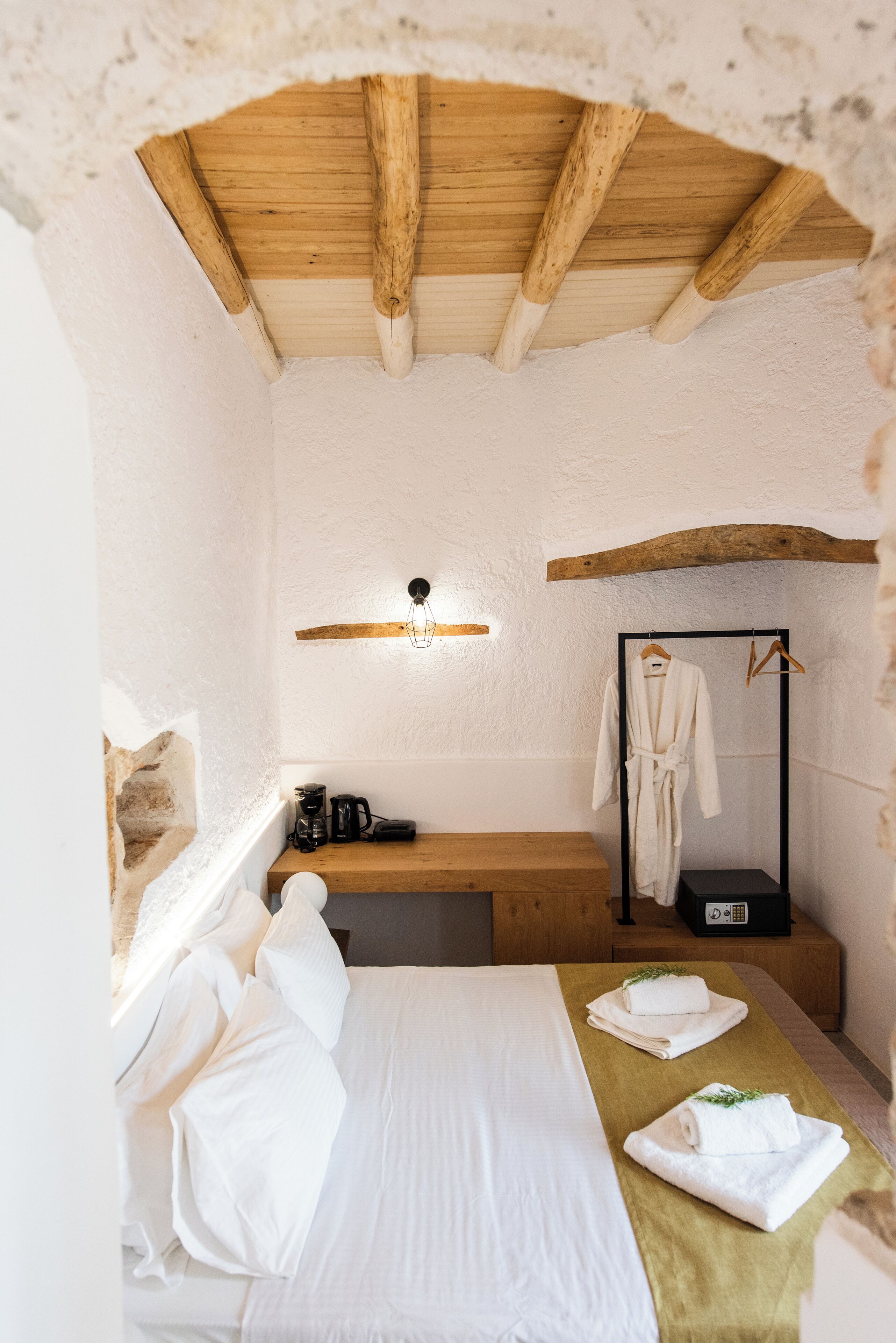 A small, rustic room with a wooden ceiling, a bed with white linens, a wooden bench, a black radio, and a robe hanging on a rack.