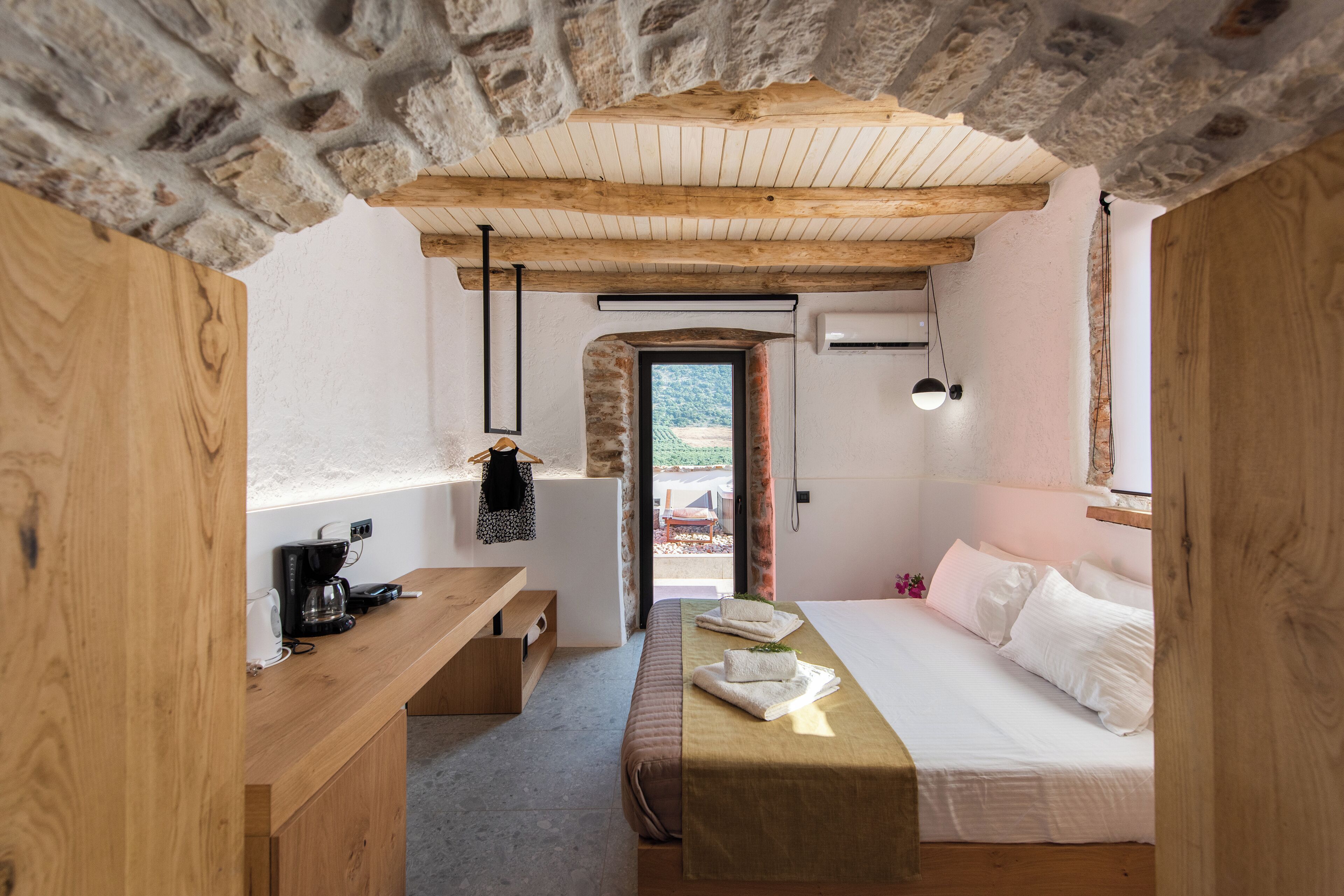 Une chambre avec un plafond en pierre, des poutres en bois, un lit recouvert de draps blancs, une table de chevet avec une machine à café, et une vue sur l’extérieur par une porte ouverte.