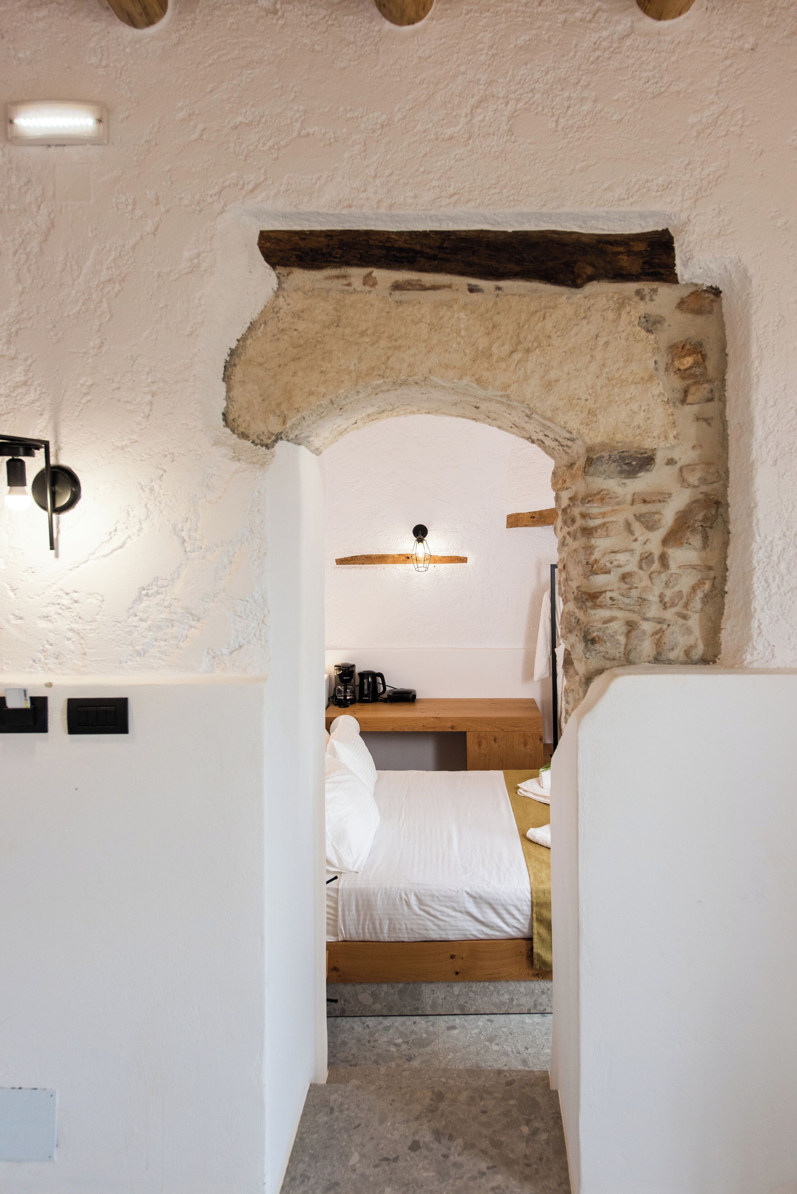 Un couloir étroit mène à une chambre dotée d’une arche en pierre, d’un lit recouvert de linge blanc, d’un banc en bois et d’une machine à café posée sur une étagère.