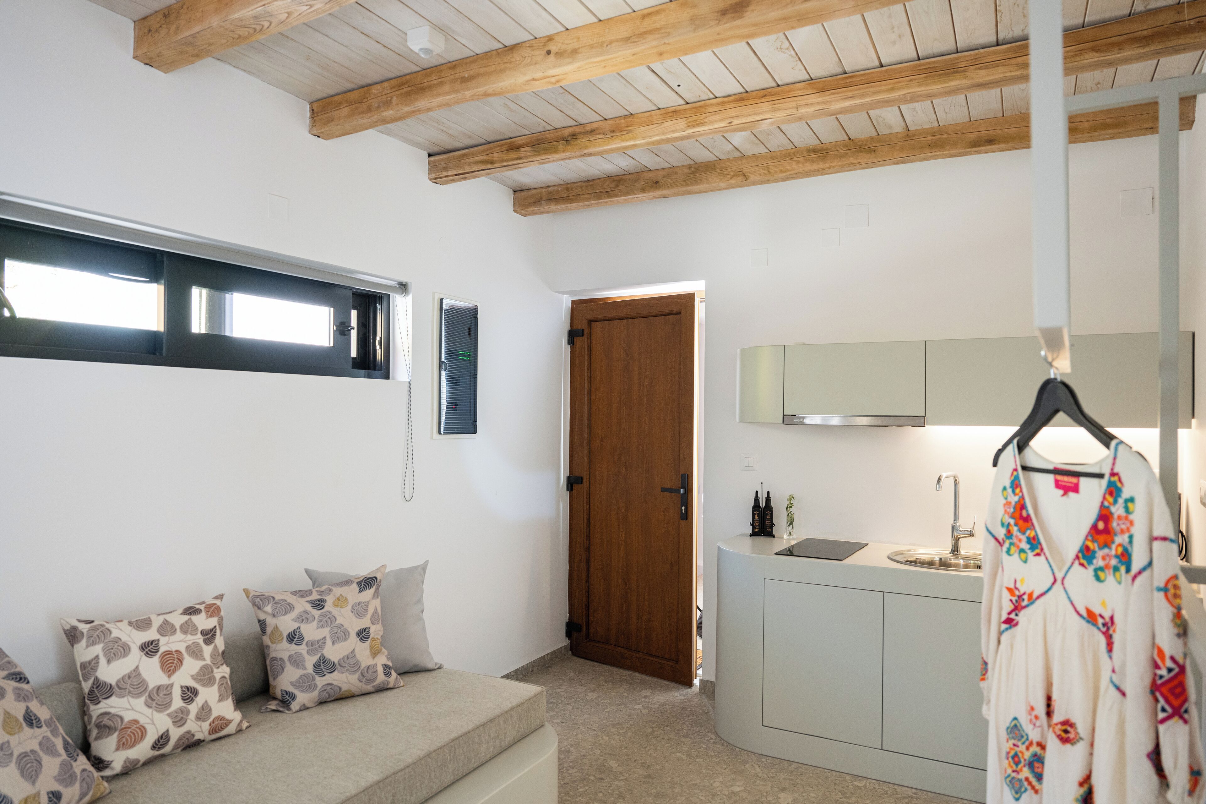 Um interior moderno com teto de madeira, sofá com almofadas estampadas, área de cozinha com pia e armários, e uma porta que dá para outro quarto.