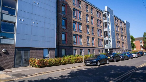 Un bâtiment moderne à plusieurs étages, avec une façade combinant briques et verre, un parking où sont garées plusieurs voitures, et un ciel d’un bleu éclatant.