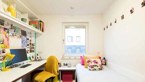 Une petite pièce bien éclairée, avec un bureau, une chaise et un lit. Une fenêtre donne sur les bâtiments situés à l’extérieur.