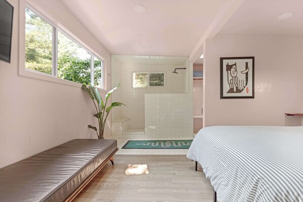 Un dormitorio con cama, un banco, planta en maceta, zona de ducha e imagen enmarcada en la pared.