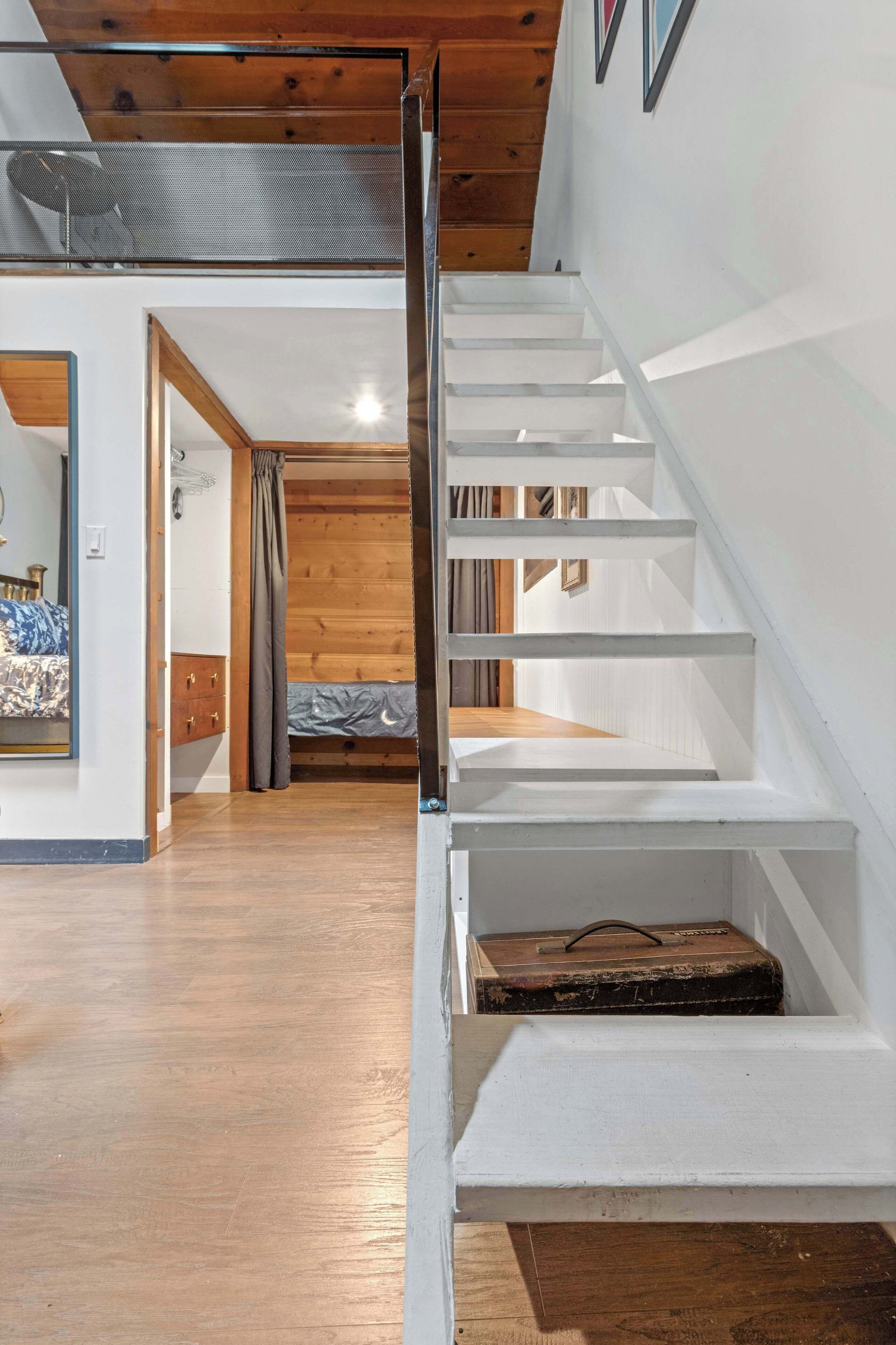 Un escalier mène à une chambre située en mezzanine, avec un plancher en bois et un lit recouvert d’une couvre-lit à motifs bleus et blancs.