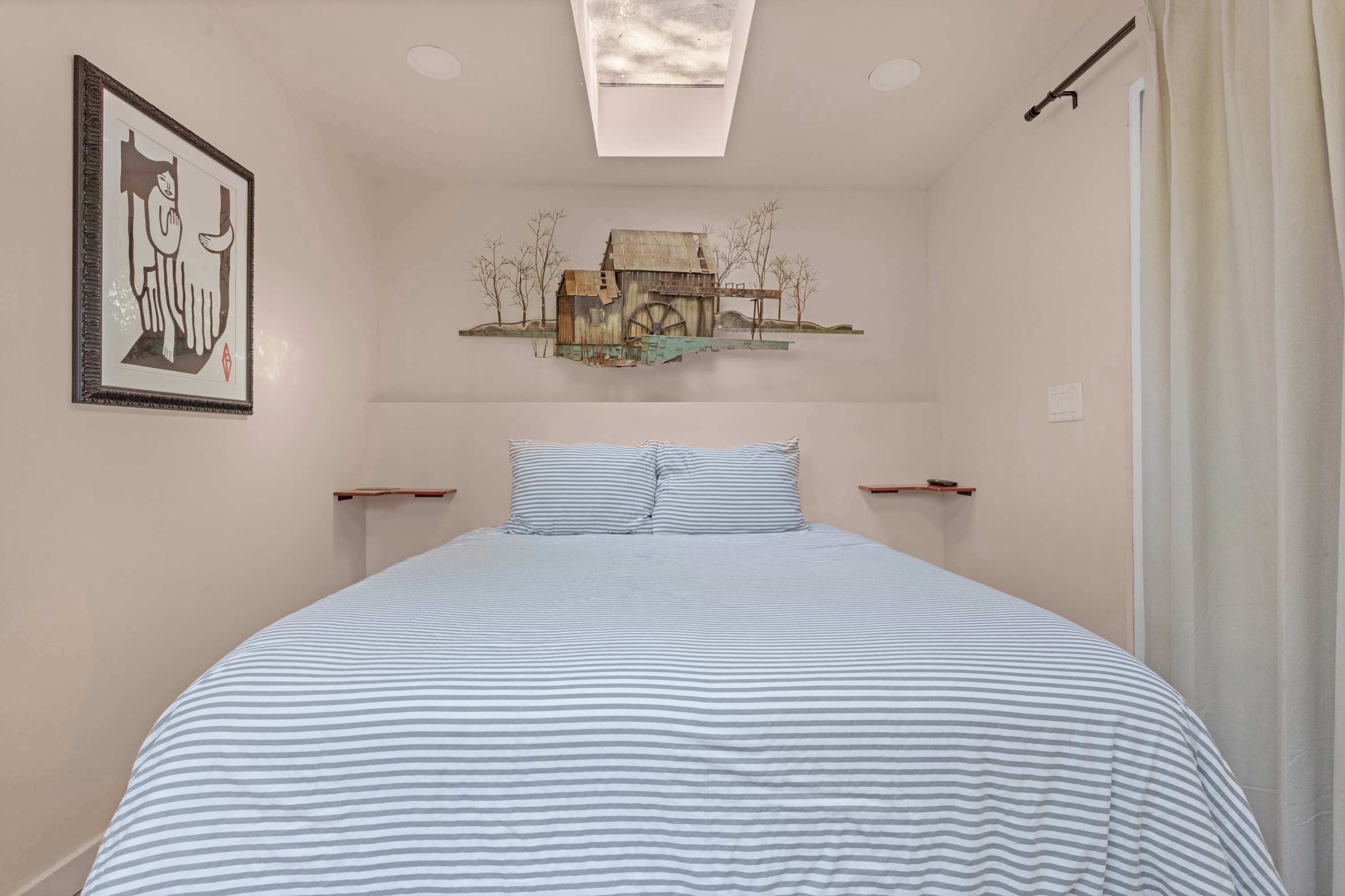Een slaapkamer met een bed, een lijstwerk kunstwerk, een plafondlamp en een wandplank met een schilderij van een rustieke watermolen.