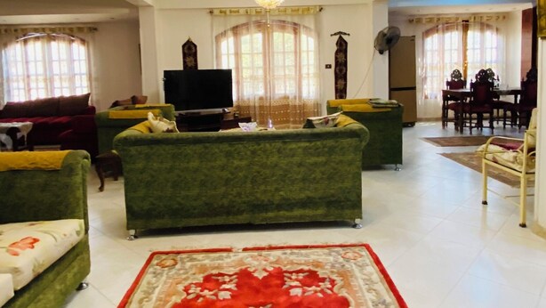A spacious living room with a large red carpet, green sofas, a chandelier, and a dining area in the background.