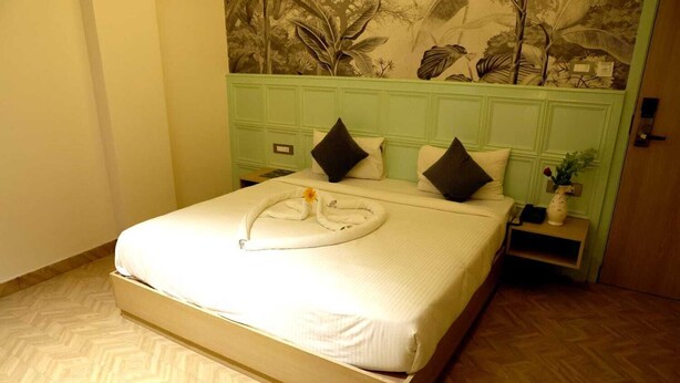 A neatly made bed with a white comforter and pillows, a small bedside table with a vase, and a wall with a tropical mural. - Sampanna Inn (Varanasi)