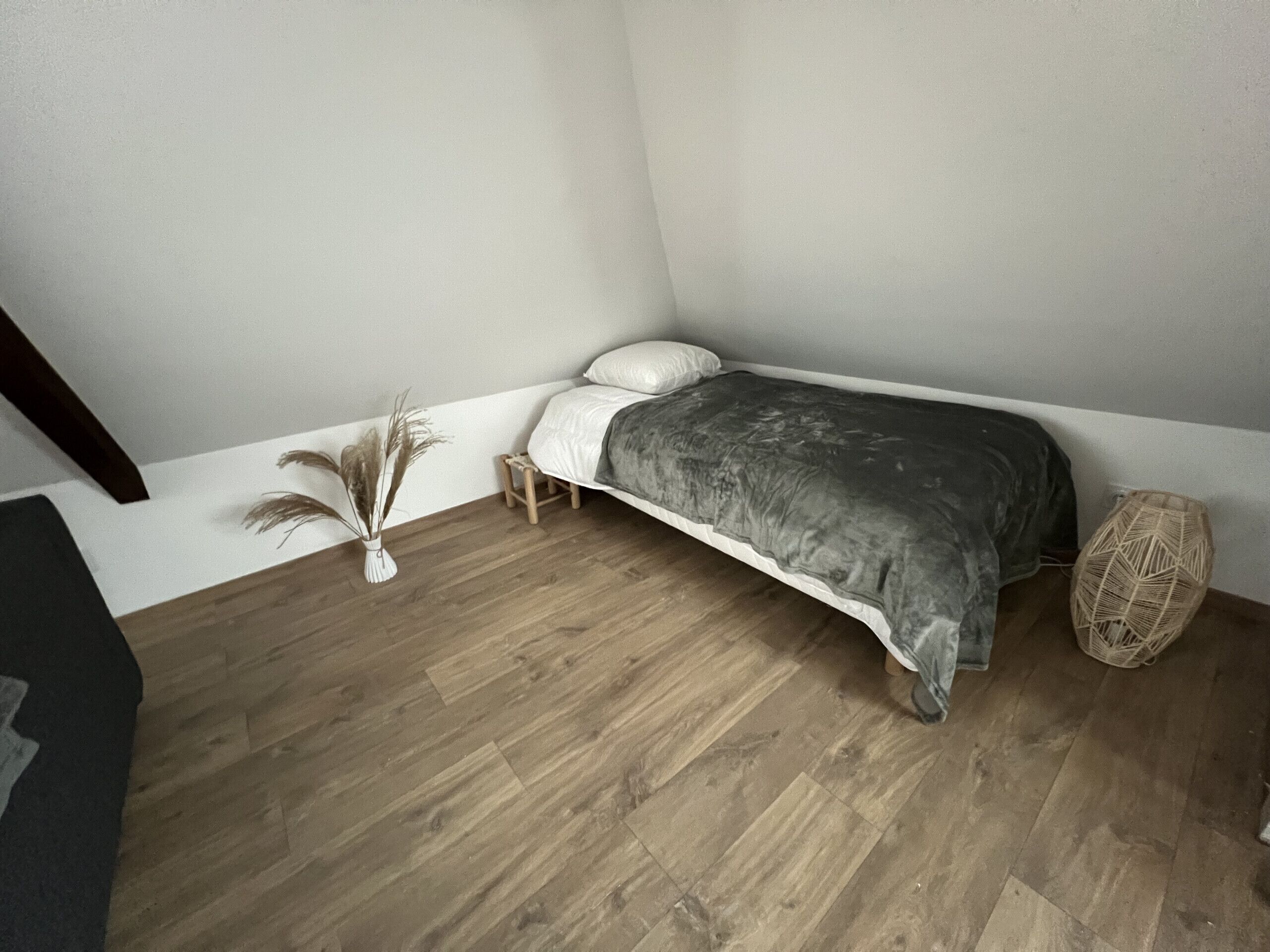 A bedroom with a bed, a small wooden stool, a vase with dried plants, and a woven basket.