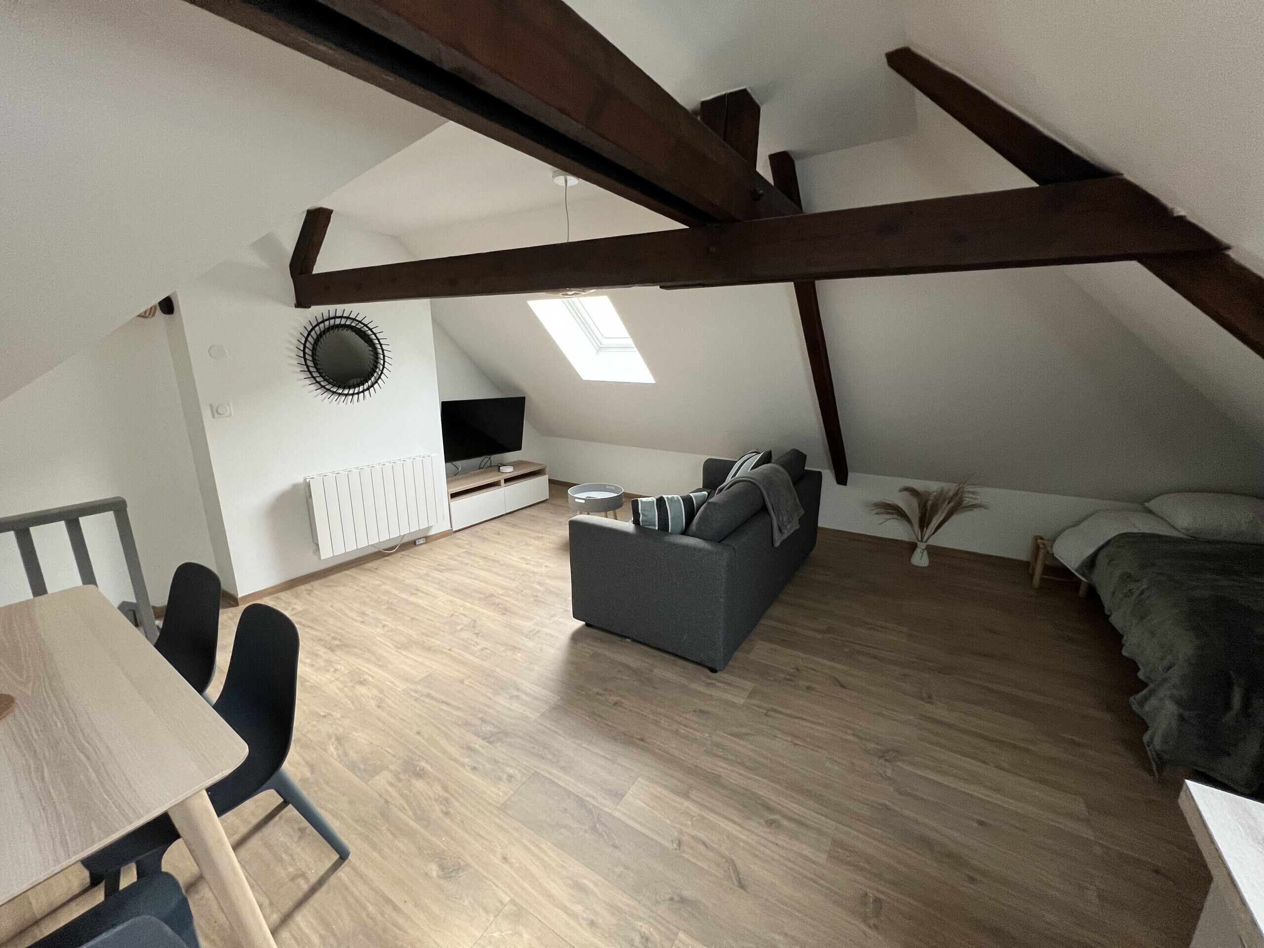 Ein Schlafzimmer im Dachgeschoss mit schrägen Wänden, einem eingebauten Fernseher, einem grauen Sofa, einem kleinen Tisch und einem Bett mit grauer Decke.