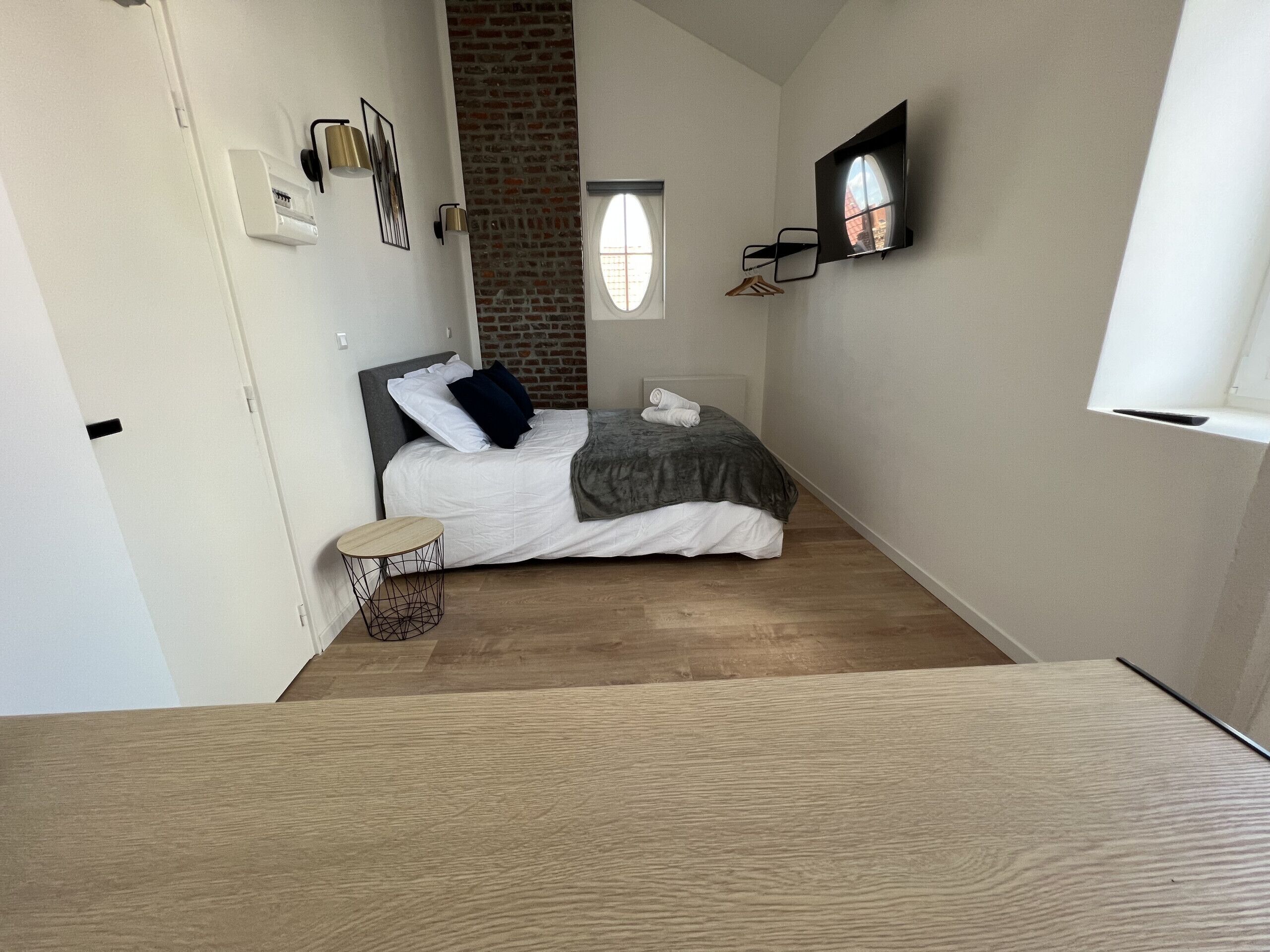A bedroom with a bed, a small round table, a hanging coat rack, and a brick wall.