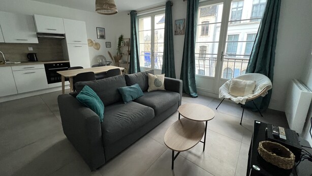 A modern apartment with a grey sofa, a small round table, a kitchen area, and large windows with curtains. - A 2 pas du Beffroi, 2-4p Wifi, Parking (Boulogne-sur-Mer)