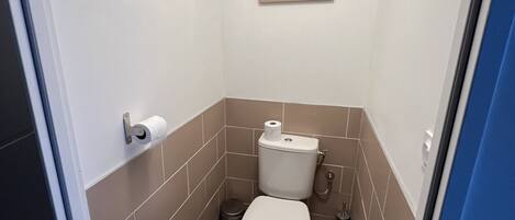 A small, compact bathroom with a toilet, a trash bin, and a wall-mounted toilet paper holder.
