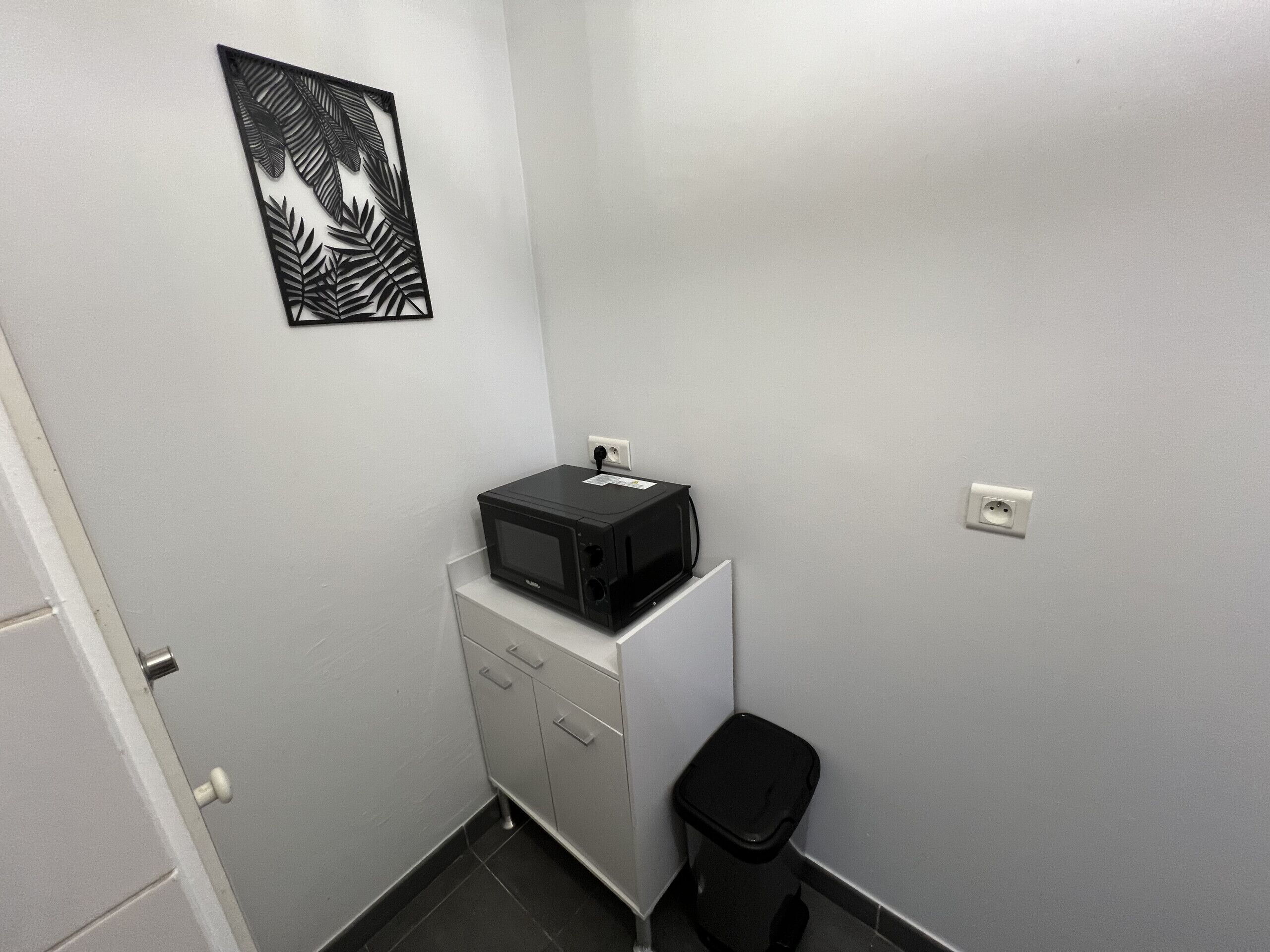 Une petite chambre d’hôtel étroite, avec un four à micro-ondes posé sur un meuble, une poubelle et une œuvre d’art accrochée au mur.
