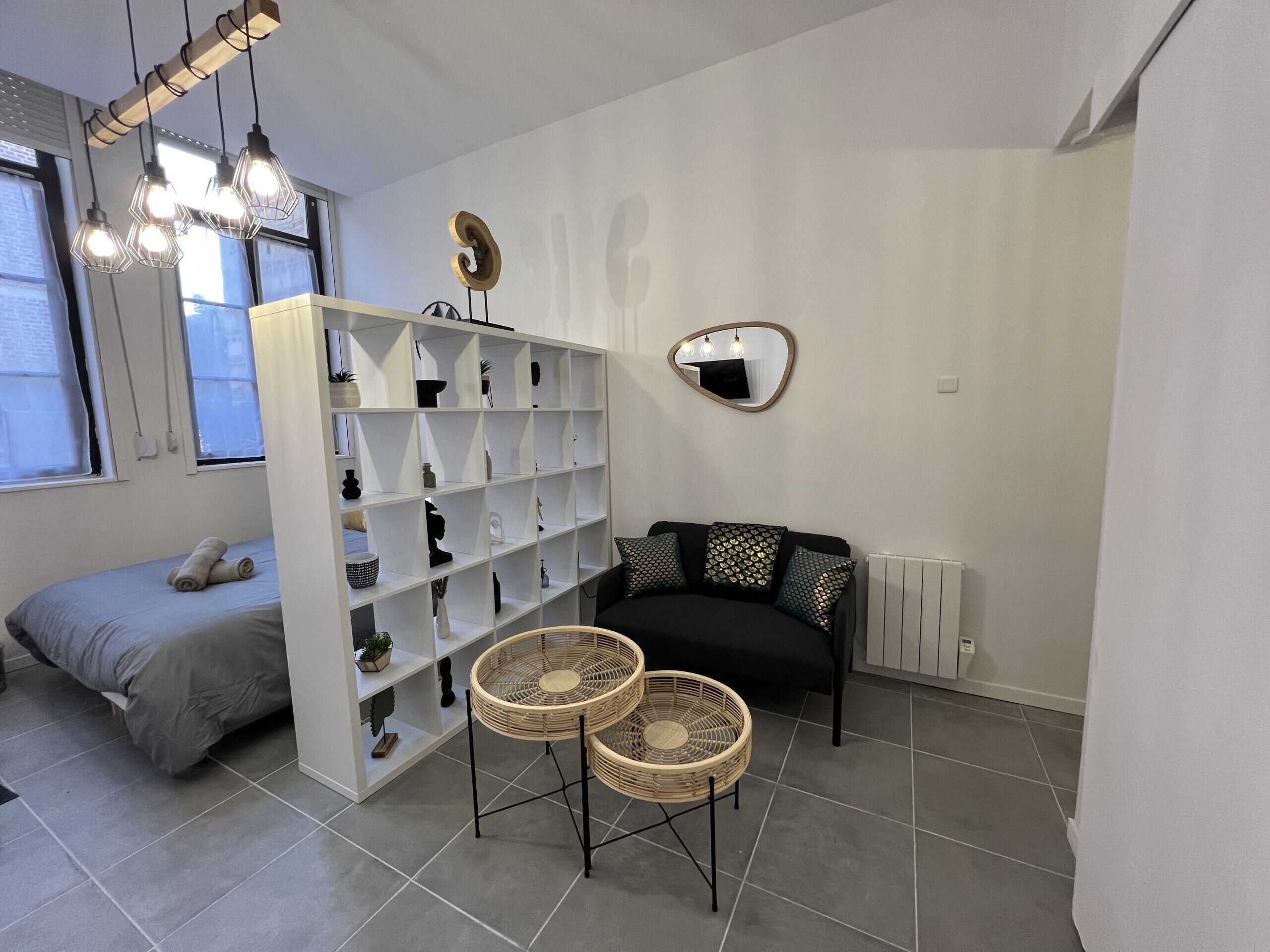 Ein modernes Zimmer mit einem Bett, einer weißen Regalkonstruktion, zwei Korbstühlen, einem schwarzen Sofa und einem Spiegel an der Wand.