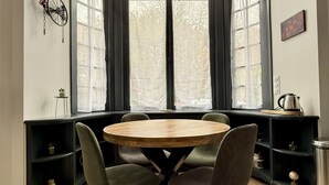 A dining area with a round wooden table and four chairs, large arched windows, and built-in shelving with various items. - Le Calais Wilson 1 : 4 pers (Calais)