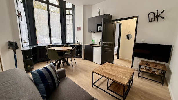A modern living space with a sofa, a wooden coffee table, a dining table with chairs, a kitchen area with cabinets, a TV mounted on the wall, and a large window with curtains. - Le Calais Wilson 1 : 4 pers (Calais)