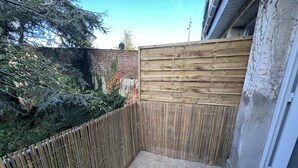 A narrow outdoor space with a wooden fence, a concrete floor, and a brick wall with vegetation.