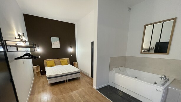 A modern bedroom with a bed, bedside tables, a hanging rack, and a large bathroom with a bathtub and mirror.