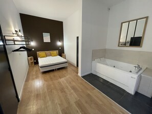 A modern bedroom with a bed, bedside tables, a hanging rack, and a large bathroom with a bathtub and mirror. - Calais Wilson 2 : Studio Balnéo 2p (Calais)