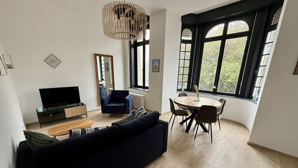 Ein modernes Wohnzimmer mit einem dunkelblauen Sofa, einem Fernseher, einem Esstisch mit Stühlen und großen Fenstern mit Blick ins Grüne.