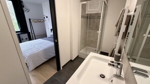Ein modernes Badezimmer mit einem großen Waschbecken, einer Duschkabine aus Glas und einem durch eine offene Tür einsehbaren Bett.