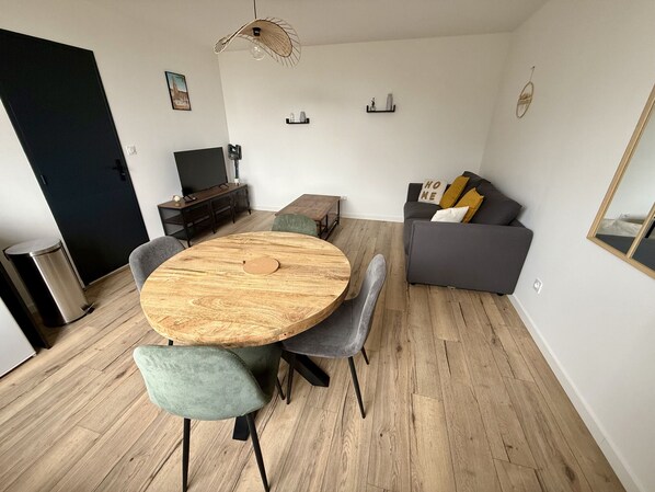 A modern living room with a round wooden dining table, grey upholstered chairs, a grey sofa with cushions, a black TV stand, and a unique ceiling fan.