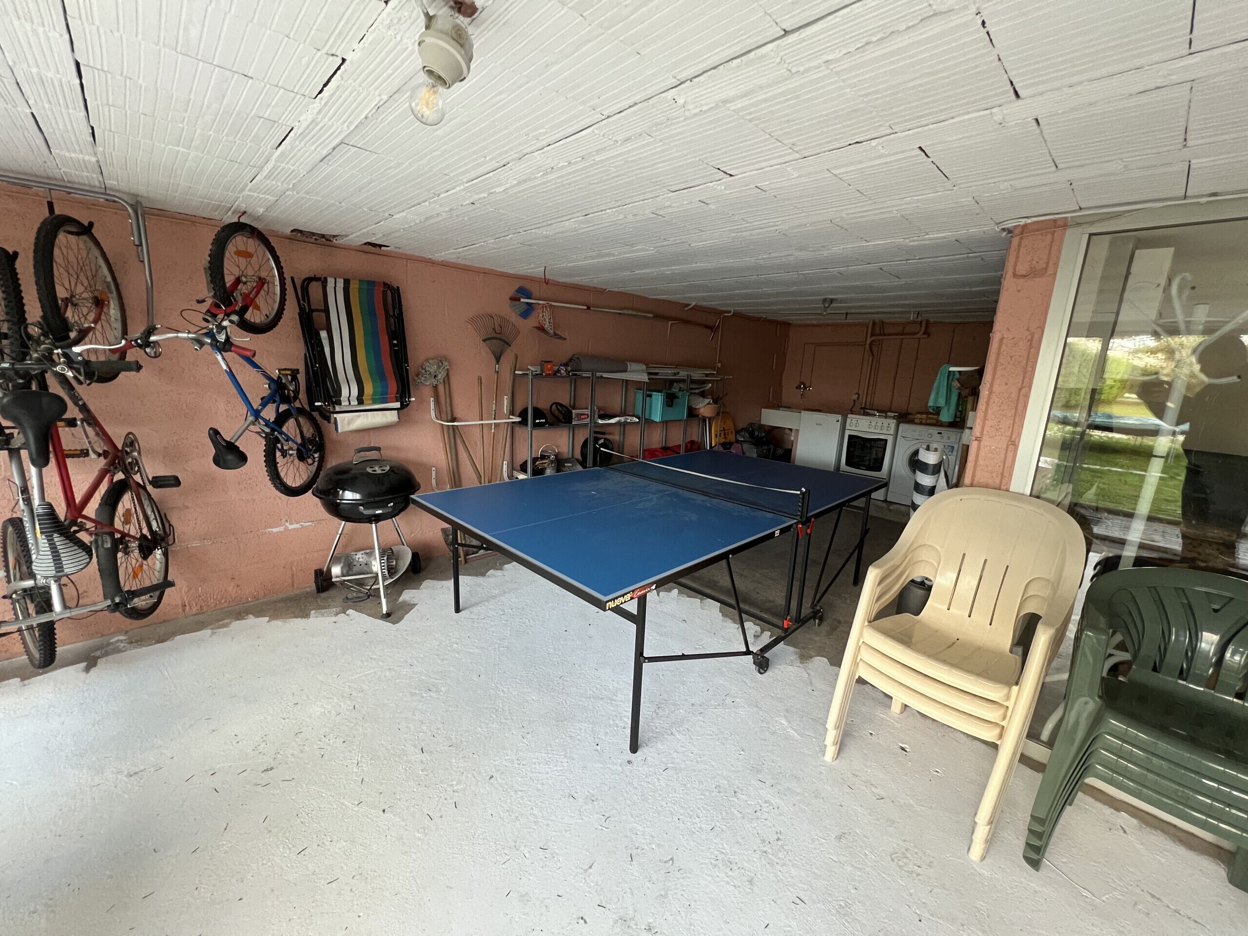 Een kamer met een pingpongtafel, fietsen, een barbecue en een wasmachine.