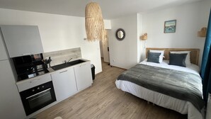 Ein modernes Hotelzimmer mit Küchenzeile, einem Bett mit grauer und weißer Tagesdecke, einer Pendelleuchte und einem an der Wand befestigten Spiegel.