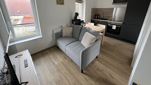 Un espace de vie compact comprenant un canapé gris, une petite table à manger, une kitchenette équipée d’appareils modernes et une fenêtre donnant sur un immeuble au toit rouge.