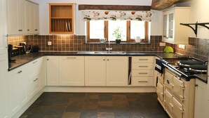 A kitchen with white cabinets, a black countertop, a stove, and a window with a floral valance. - Beachside holiday home  (Padstow)