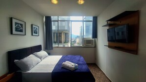 A hotel room with a bed, a TV on the wall, and a window with a city view. - Seu aconchegante lar no centro carioca (Rio de Janeiro)