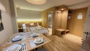 Ein modernes Hotelzimmer mit Holzboden, einem Bett mit Kopfteil, einer Duschkabine und einem Esstisch für zwei Personen.