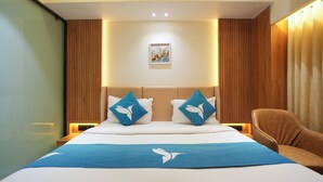 Habitación de hotel con cama, dos almohadas, mesita de noche, una silla e imagen colgada en la pared.